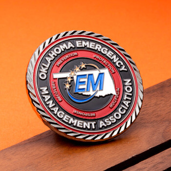 Circular Oklahoma Emergency Management Association challenge coin with a red and black border, silver rope edge, and blue EM logo over an Oklahoma state map.