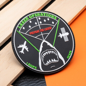 Circular PVC patch for Laser Operations featuring a shark with green laser beams, a satellite, and an airplane on a black background.