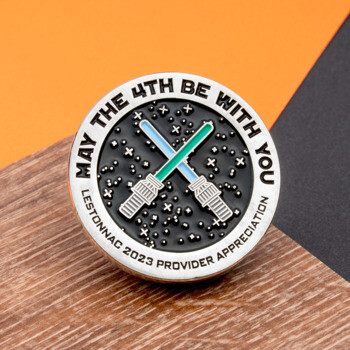 Circular challenge coin featuring crossed lightsabers on a black starry background with the text May the 4th Be With You and Lestonnac 2023 Provider Appreciatio