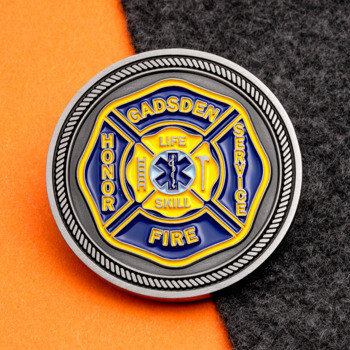 Circular Gadsden Fire and EMS challenge coin with a blue and yellow Maltese cross, Star of Life, and silver rope edge, set on orange and grey background.