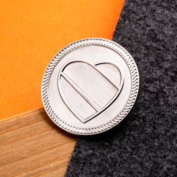 Circular silver lapel pin featuring a stylized heart emblem and a classic rope border on a multi-textured background.