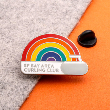 SF Bay Area Curling Club enamel lapel pin shaped like a curling stone with a colorful rainbow arch on a split grey felt and orange background.