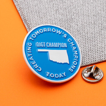 Round blue lapel pin featuring the shape of Oklahoma and OJGT Champion text, shown with a metal butterfly clutch.