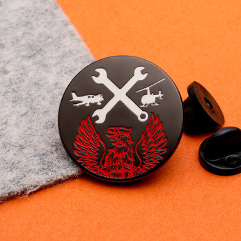 Circular black enamel lapel pin with crossed wrenches, an airplane, helicopter, and a red eagle emblem.