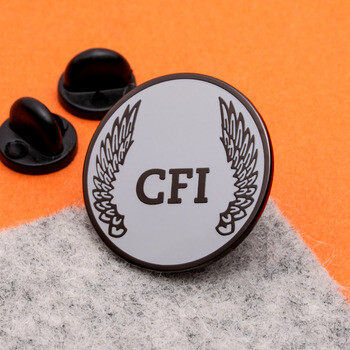 Circular lapel pin with black trim, silver background, CFI lettering, and wing illustrations, shown with rubber clutches on orange and gray felt.